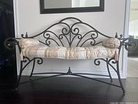 Full front view of the wrought iron bench with patterned metalwork and cream padded seat cushion with bolsters on each side.