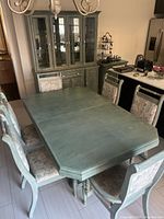 Full view of the dining table set with 8 upholstered chairs, showing the dining table with leaf extension, painted oak wood in muted green/gray and chairs with floral upholstery.