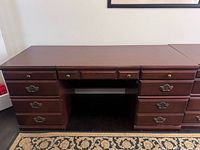 Photo showing dark brown wooden desk with three drawers in the middle and two columns of drawers on either side, placed on a decorative rug.