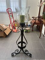 Front view of wrought iron candle holder stand with etched glass hurricane shade and pillar candle inside, displayed on tiled floor with chairs and boxes in background.