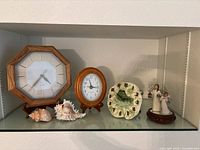 Shelf showing two wood-framed clocks, frog wall clock, two sea shells and two angel figurines on mirrored base