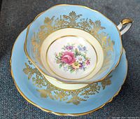 Top view of teacup nested in saucer showing blue ground, gold gilt scrollwork, floral centre