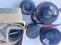 Top view of four lenses: Spiratone fish-eye lens, Minolta 50 mm lens, Pentax lens with cap, Chinon lens boxed with cap