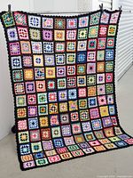 Full view of crocheted tablecloth hung vertically showing colorful granny square pattern with black borders.