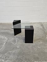 Side table shown diagonally with clear thick glass square top and two black painted wooden legs: one cube-shaped and one prism-shaped with slant edges, on concrete floor against a white wall.
