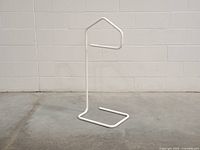 Front angle view of the white metal valet stand with geometric hanger top and rectangular base on concrete floor.