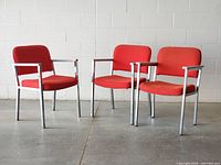 Front and side angle of the three vintage red fabric upholstered armchairs with chrome metal frames and white wood armrests in a concrete floor setting.