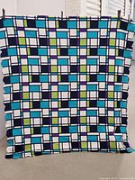 Full view of the throw blanket hanging, showing the geometric blue, white, green, and turquoise plaid pattern with black grid lines.