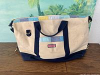 Full view of duffel showing natural canvas body, navy handles and base, multicolor whale-pattern strip and gold zipper