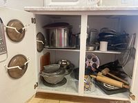 Cabinet interior displaying assorted cookware, mixing bowls, strainers, wooden mallet and rolling pin, plus glass lids mounted on door
