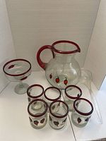 Glass pitcher with red handle and rim, five glass tumblers, one stemmed glass, and plastic ladle displayed in white background. Shows all items in the set and chili pepper design.