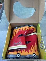 Pair of bright red children's rain boots with flame designs and fire truck wheel accents inside an open box.