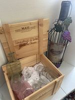Wooden MASI Agricola crate with open lid showing glass cheese trays shaped like wine bottles and multiple clear drinking glasses.
