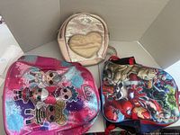 Three kids backpacks displayed side by side. One LOL Surprise-themed with character graphics, one Marvel Avengers-themed with superhero images, and one smaller beige backpack with a sequin heart design.