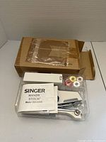 Photo showing boxed contents including handheld sewing machine, instruction manual, multiple thread spools, and original box.