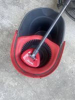 Top-down view of black and red mop bucket with spin wringer, mop handle inserted with red triangular mop head attached.