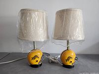 Full view of both CFL Hamilton Tiger-Cats helmet table lamps with beige fabric shades covered in plastic wrap.