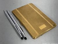 Two silver-tone Cross pens placed beside closed gold-tone textured cigarette/card case.