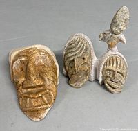 Front view of two indigenous art stone face carving sculptures on a gray surface, showing their detailed facial features and bird figure on one sculpture.