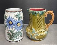 Front view of two pottery items: a yellow-green ornate pitcher with an orange handle and a white floral stein with blue flowers.