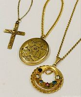 Three vintage yellow gold filled necklaces displayed side by side: cross pendant, oval floral locket, round colored gemstone pendant
