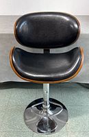 Front view of the barstool showing black cushioned seat and backrest with curved wooden framing, chrome pedestal base with footrest.