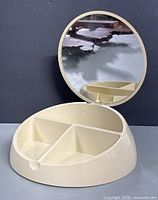 Open beauty box with round mirror and three cream-colored plastic compartments inside.