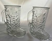 Front view of both clear glass boot-shaped mugs showing ornate pattern and handles