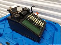 Front left view of Burroughs adding machine on blue bin showing keyboard, paper roll, ribbon spools
