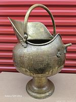 Front view of brass scuttle showing pedestal base, bail handle, spout