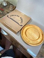 Cardboard box labeled 'Adam’s Cheese Wheel' with a graphic of cheese wedge and handle, alongside the wooden cheese wheel placed on a gray surface