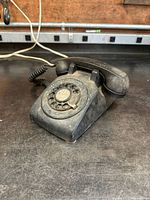 Front angled view of black rotary dial phone showing rotary dial, handset, and coiled cord on a workbench surface.