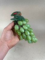 Hand holding decorative green grapes cluster against plain background showing full bunch and attached leaves.