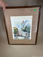 Full view of framed watercolor painting with beige mat and brown frame showing an urban street scene featuring a white building with a tower