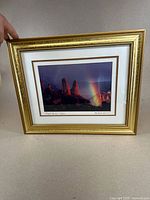Framed photograph showing the full frame and image. Gold detailed frame and mat with the photograph centered inside. Rainbow over red rock formations visible.