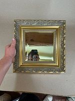 Full view of the small square vintage mirror showing the gold gilded ornate frame, held with hand for scale.