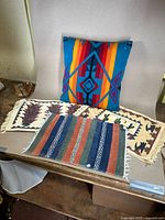Overview of multicolored pillow with geometric design and five woven placemats including cactus and pepper motifs