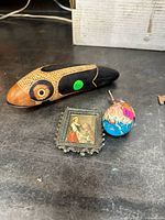 Lot items arranged together on a table: fish-shaped wooden rattle, vintage metal frame with painting, decorated egg rattle