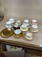 Nine fine China tea cups with matching saucers displayed on table showcasing various floral patterns and gold accents, plus two gold-tone saucers.