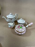 Four-piece vintage porcelain china set with teapot, sugar bowl, creamer, and handled pan with floral designs and gold accents.