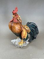 Front and side view of a large colorful rooster figure statue showing detailed feathers and base.