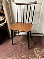 Full view of chair showing walnut seat, tapered black legs, and vertical black spindle back supports, with visible wear on flooring beneath.