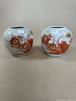 Pair of round-bodied Asian ginger jars in white ceramic with red and gold floral and bird designs sitting side by side.