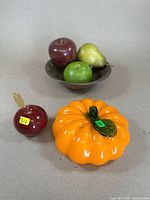 Photo showing a copper bowl containing green apple, yellow pear, and red apple with a separate Murano glass red apple and an orange glass pumpkin with green leaf and stem in the foreground