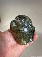 Front view of small verdite sculpture of seated man resting head on arms, polished stone with mottled green shades.