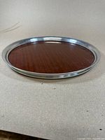 Photo showing round serving tray resting on a surface, with visible mahogany base and reflective silver-tone metal rim.