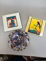 Three Indigenous American decorative art pieces displayed on a surface: two framed art tiles and a round decorative plate with feathers and beadwork attached around its edge.