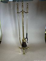 Full view of the vintage brass fireplace tool kit on its stand showing the poker, shovel, and brush suspended on the stand with a tripod foot base.
