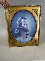 Rectangular gold frame with oval opening, showing a painted portrait of Jesus Christ with long hair and beard in profile, held vertically.