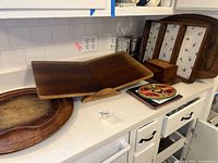 Photo of assorted wood trays, a wood cookbook stand, decorative ceramic tiles, and a small metal canister all displayed on a white countertop.